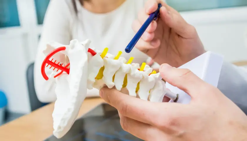 Dłoń lekarza wskazująca niebieskim długopisem na model kręgosłupa.